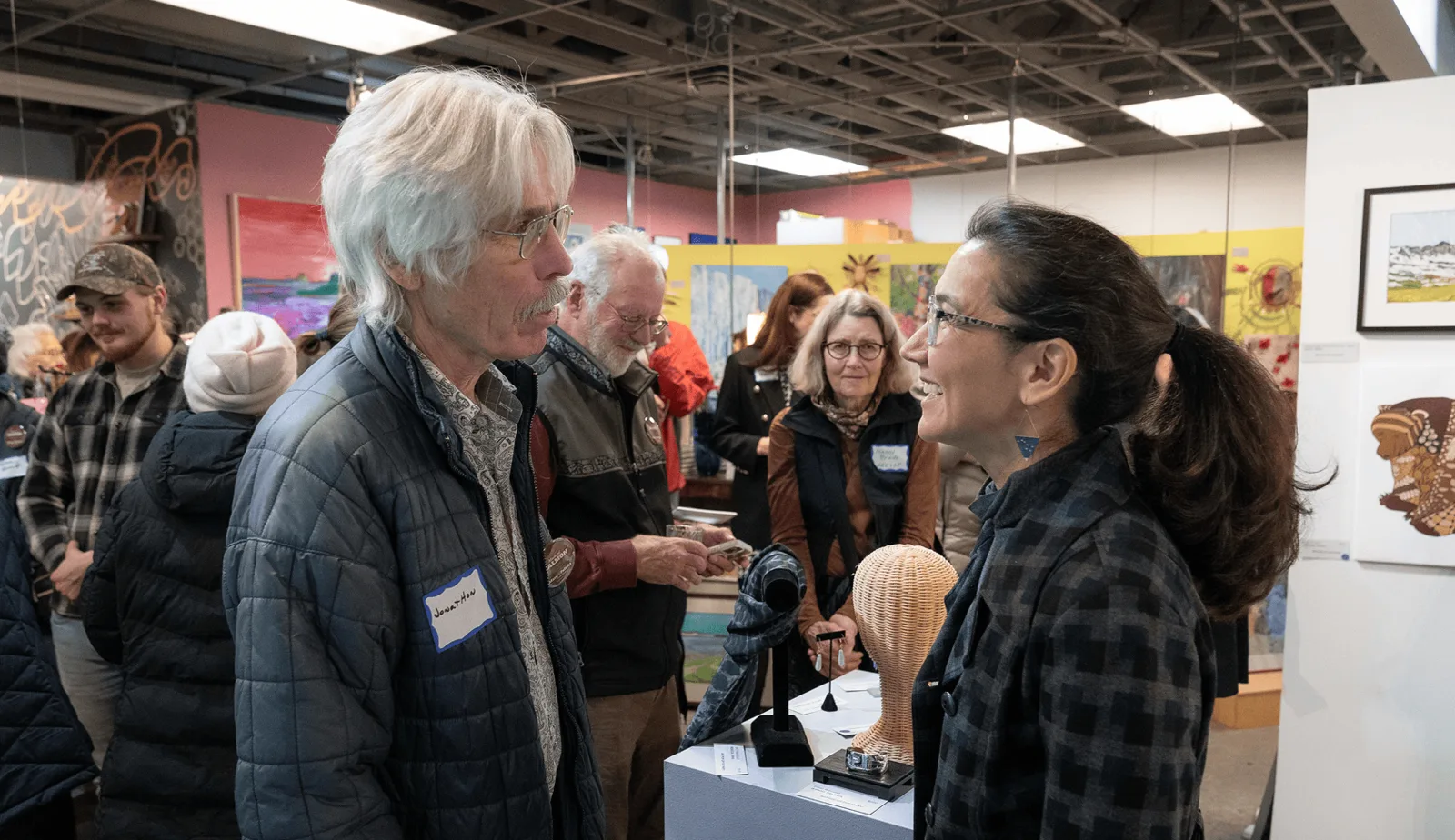 An older man with white hair speaks with Mary Peltola at an art fair booth displaying woven sculptures and jewelry.