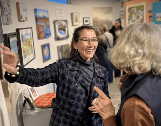 Mary Peltola laughing with a visitor among paintings at a gallery.