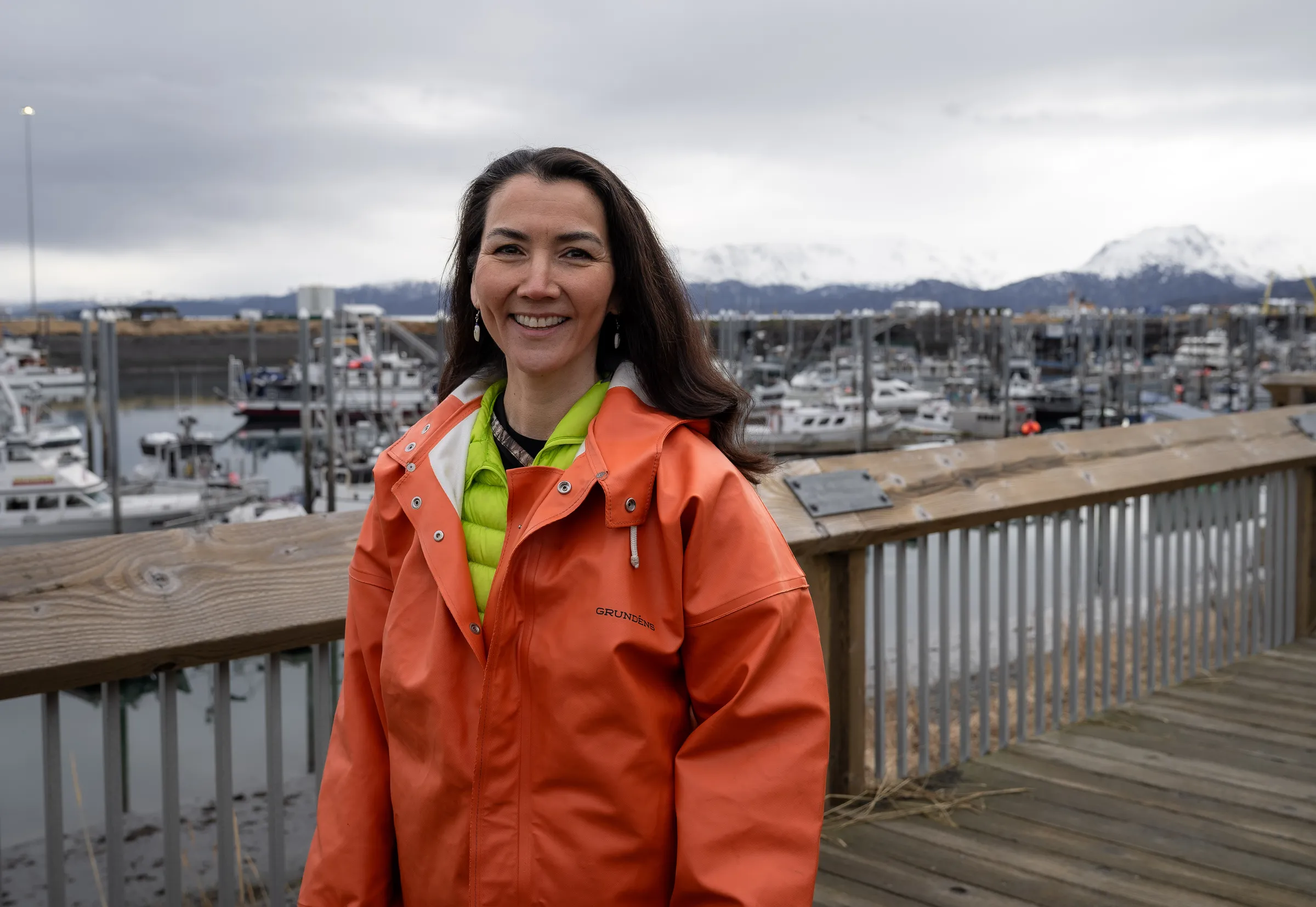 Mary Peltola in an orange rain jacket at a small-boat harbor, snowy mountains behind her.