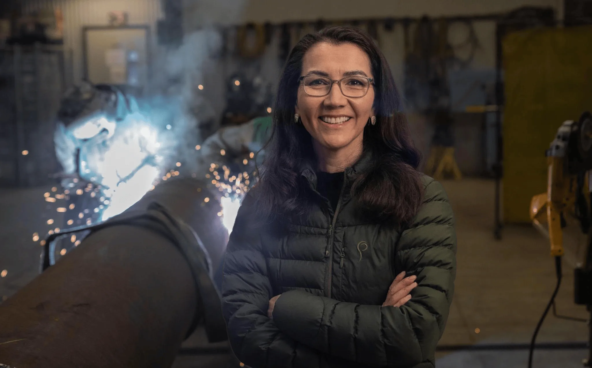 Mary Peltola in a welding shop, a welder at work beside her.