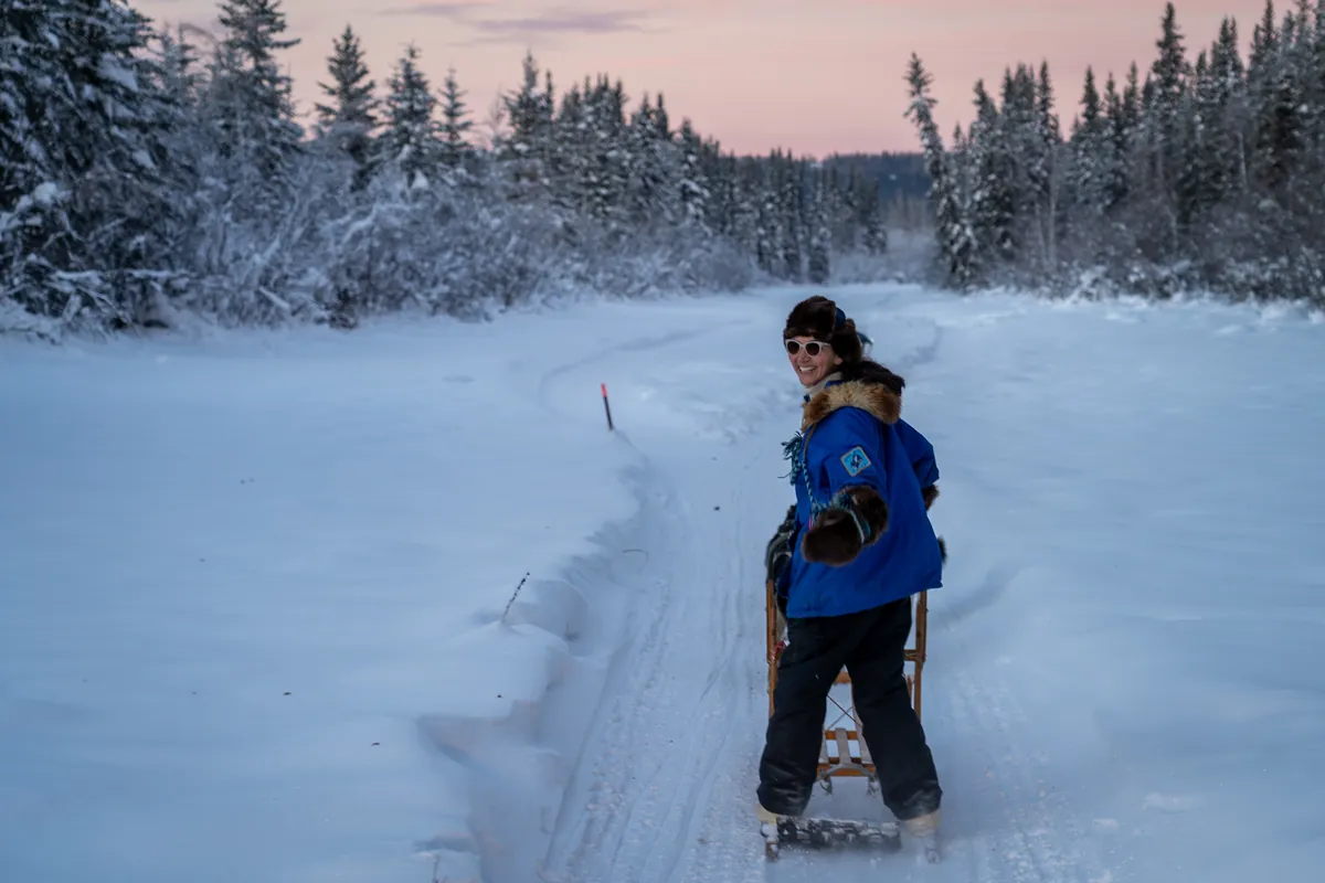 Mary Peltola looks back with a smile from her dog sled on a snowy spruce trail at dusk.