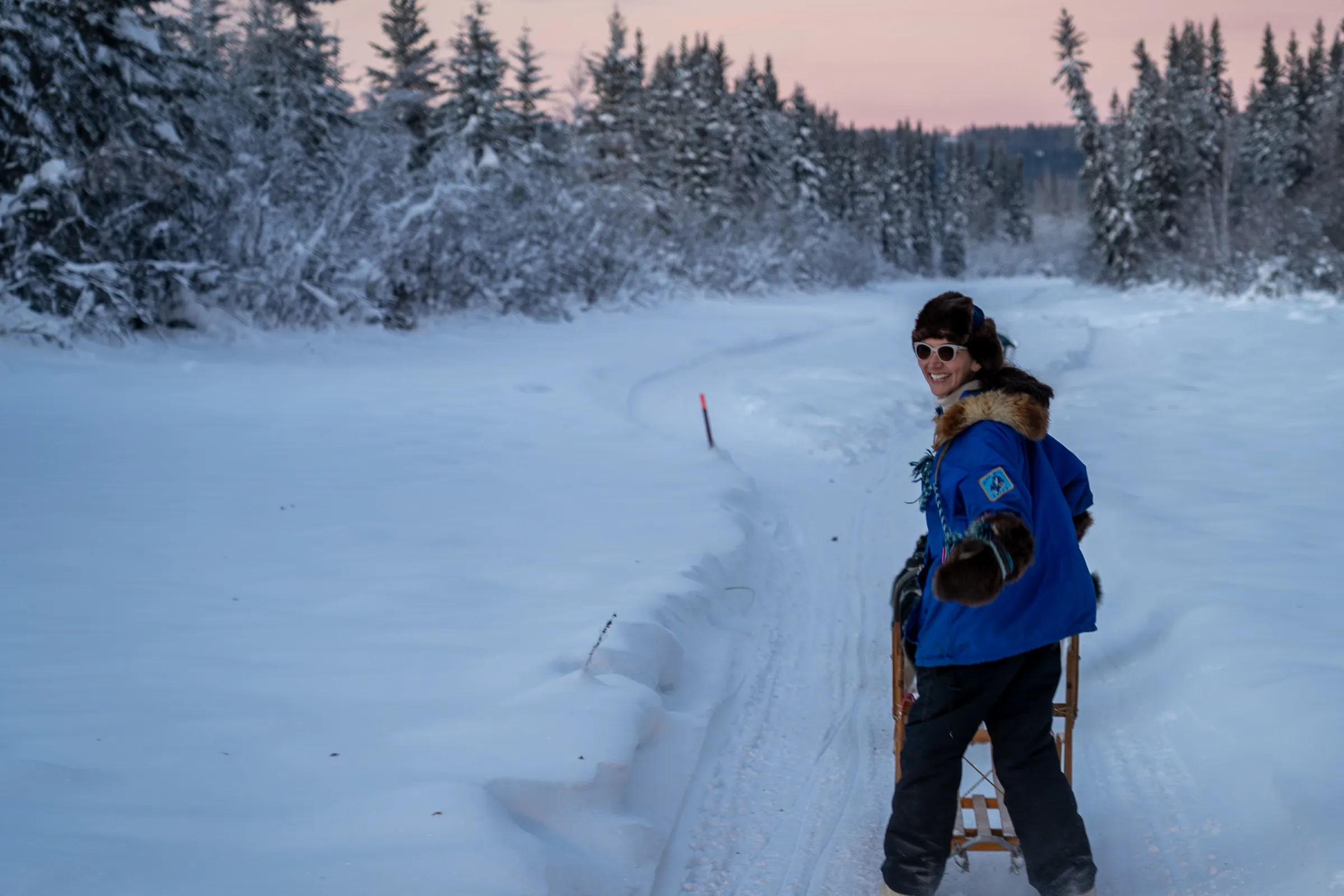 Mary Peltola looks back with a smile from her dog sled on a snowy spruce trail at dusk.