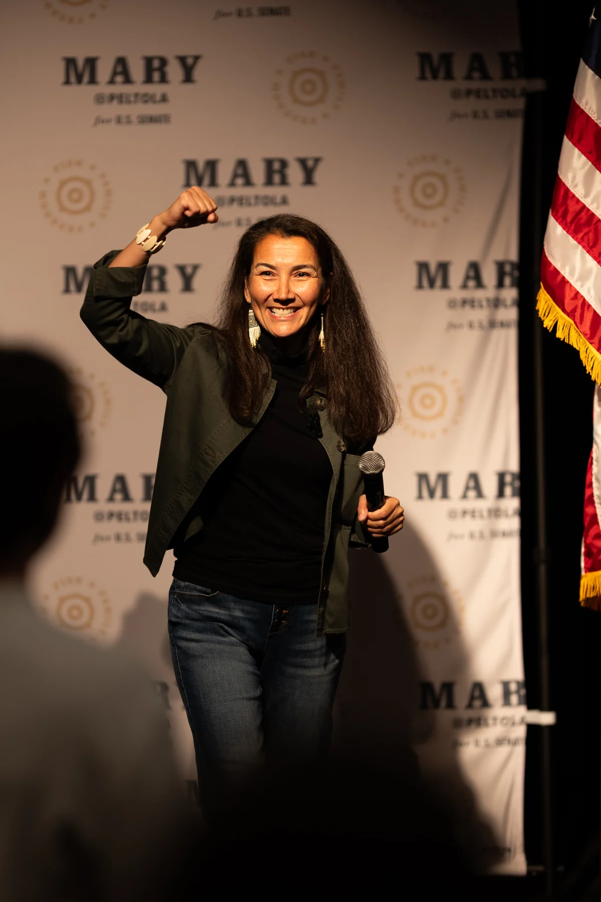 Mary Peltola on stage, fist raised, in front of her campaign backdrop and the American flag.