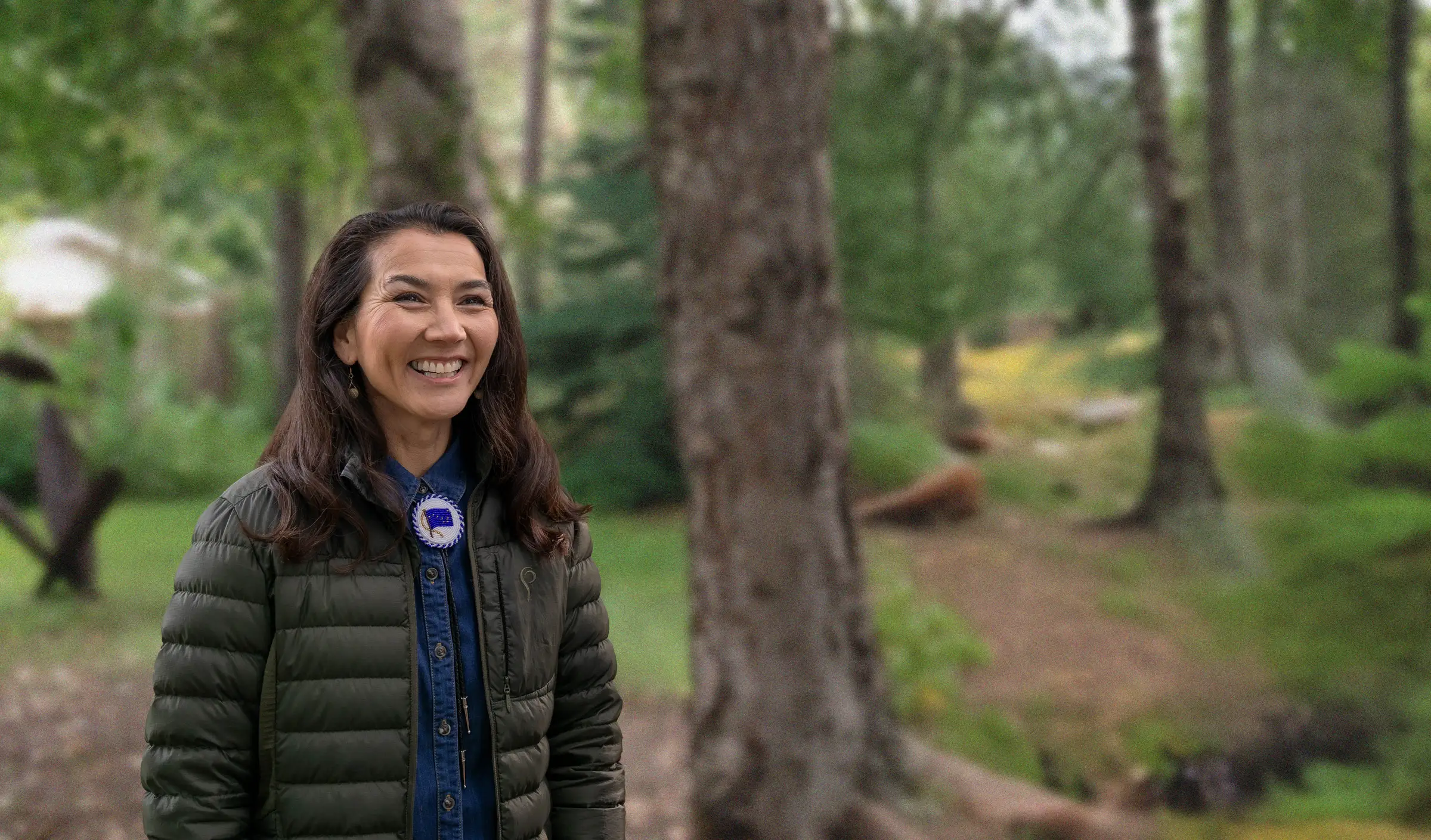 Mary Peltola smiling among spruce trees in a quilted green jacket.